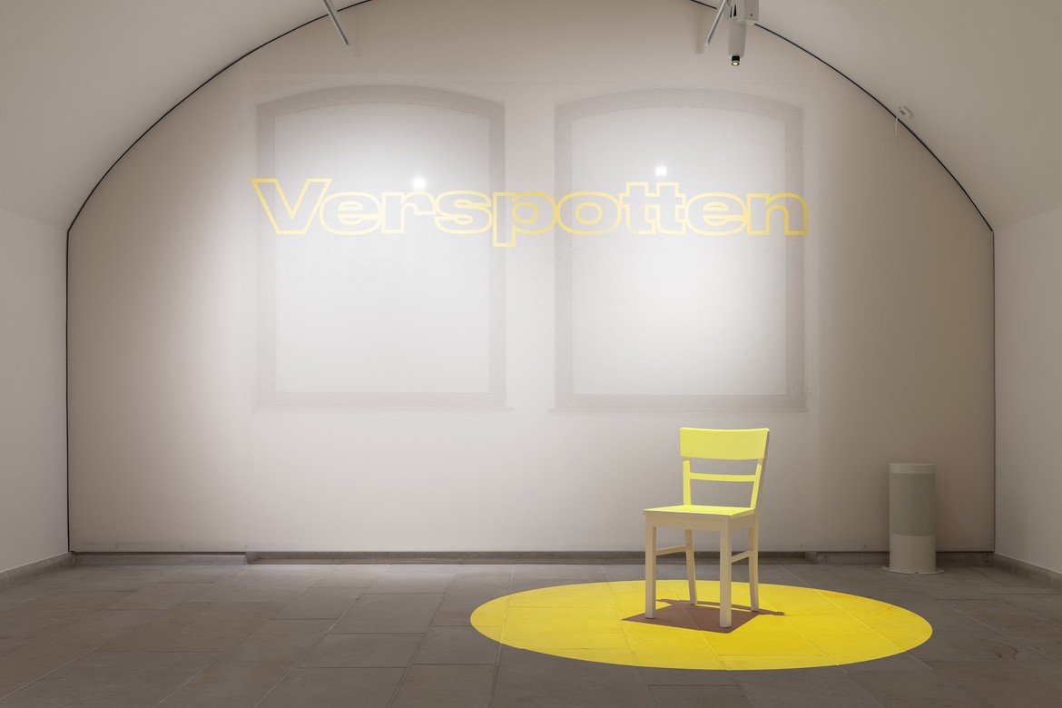 Foto: Blick in den ausstellungsraum "Spotten" mit einem kleinen weißen Stuhl in einem gelben Lichtkreis
